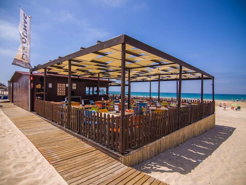 Strandbar aus Holz mit Überdachung, Tischen und Stühlen, umgeben von Sand und blauem Himmel.