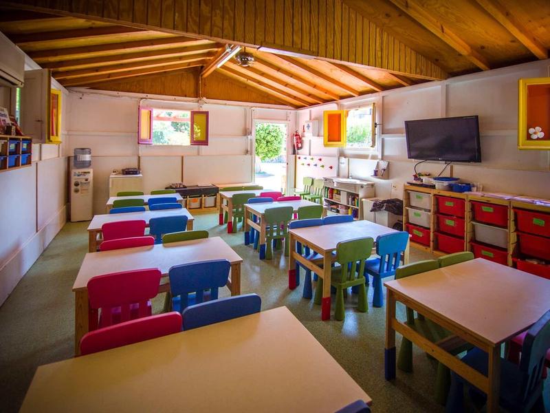 Bunt eingerichteter Klassenraum mit kleinen Tischen, Stühlen, großer Tafel und Fernseher unter Holzdach.