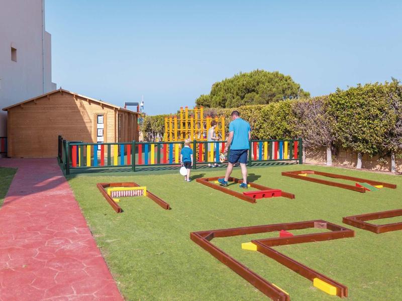 Kind spielt auf kleinem Minigolfplatz mit buntem Holzzaun und hölzernem Gartenhaus im Hintergrund.