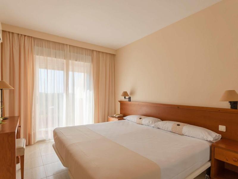 Helles Schlafzimmer mit Doppelbett, Holzmöbeln und großen Fenstern mit Gardinen.