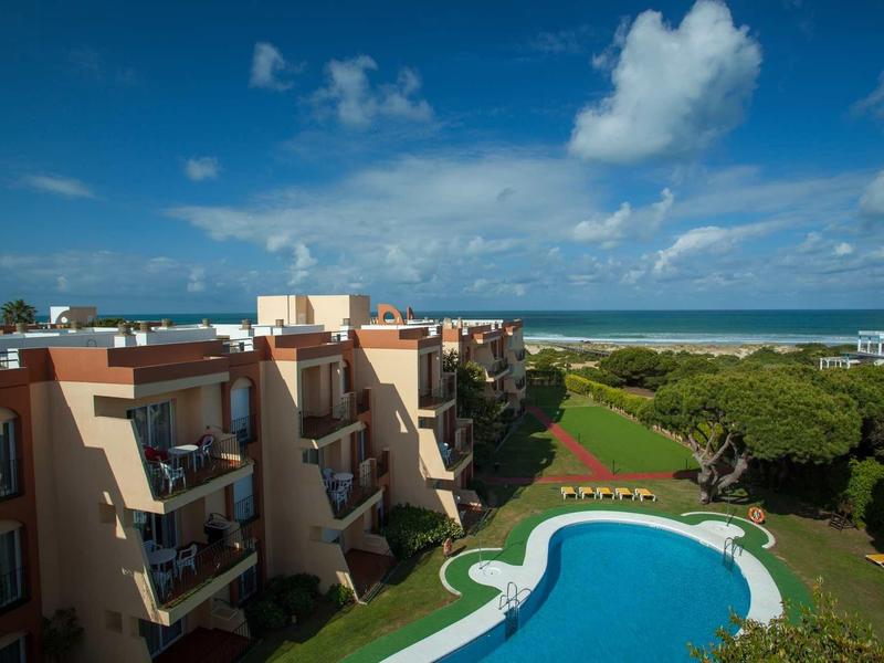 Ein großer, blauer Pool vor mehreren terrakottafarbenen Gebäuden mit Meer im Hintergrund und blauem Himmel.