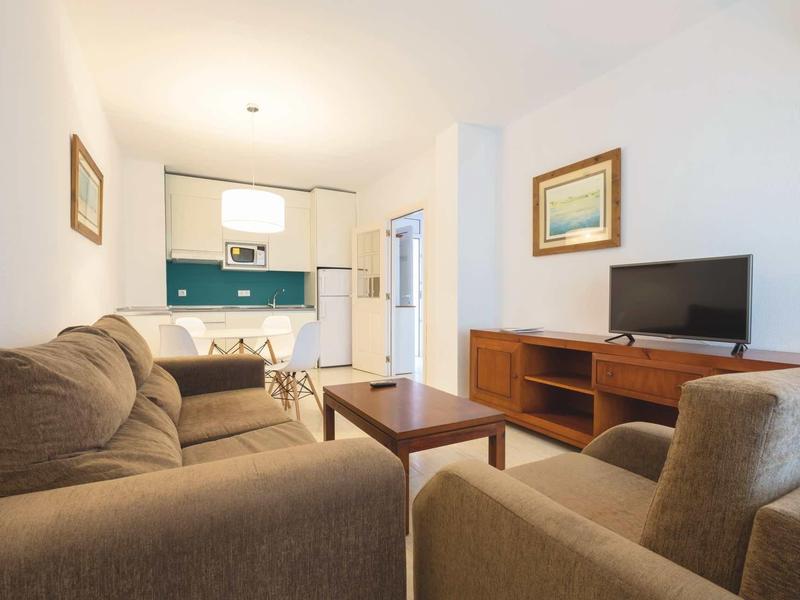 Gemütliches Wohnzimmer mit grauen Sofas, Holztisch, TV und offener Küche im Hintergrund.