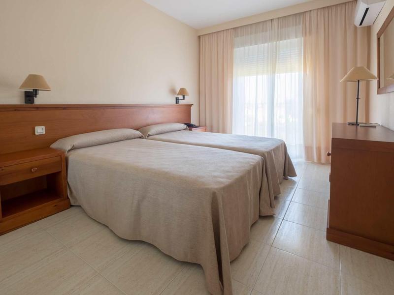 Helles Schlafzimmer mit zwei Einzelbetten, Holzmöbeln und großen Fenstern mit Vorhängen.