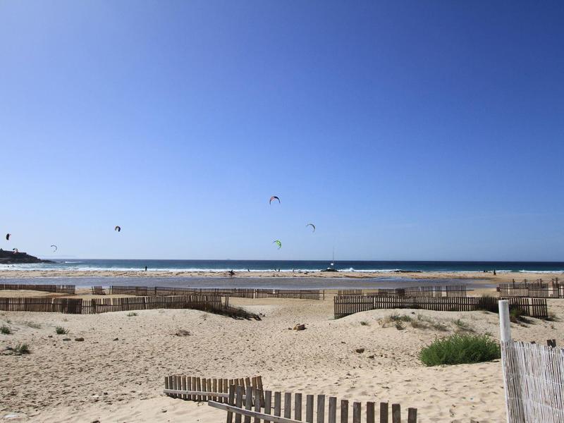 Spiaggia di sabbia con recinzioni di legno e kitesurf sotto un cielo azzurro limpido sul mare.