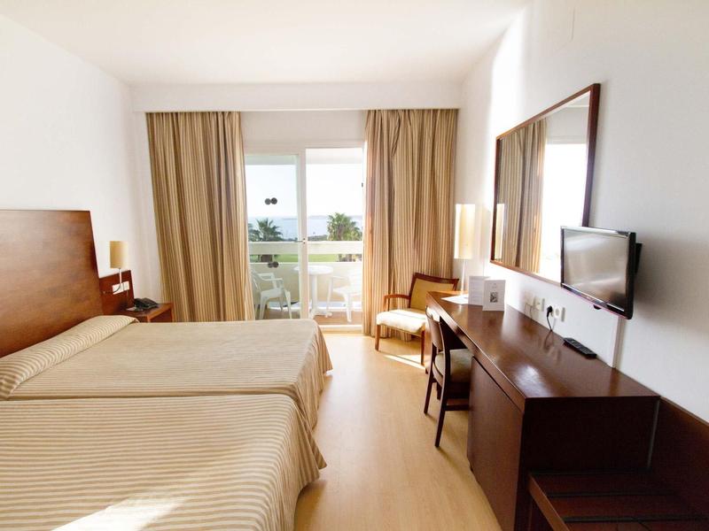 Camera d'hotel luminosa con due letti singoli, scrivania, sedia e balcone con vista mare.