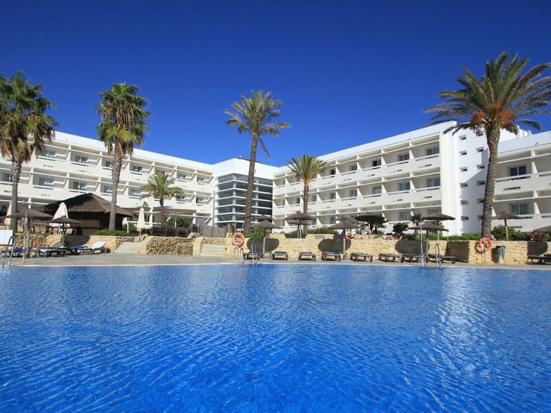 Complesso alberghiero moderno con grande piscina e palme sotto un cielo azzurro limpido.