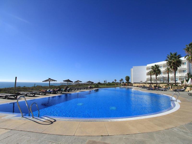 Piscina rotonda dell'hotel con lettini e palme sotto un cielo azzurro limpido.