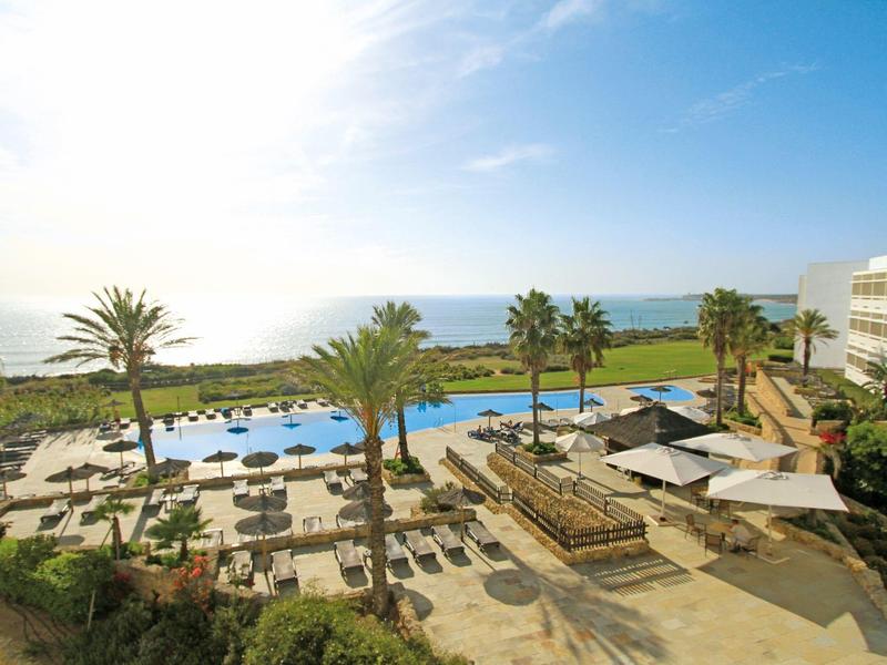 Area piscina dell'hotel con palme, lettini e vista mare sotto un cielo azzurro.