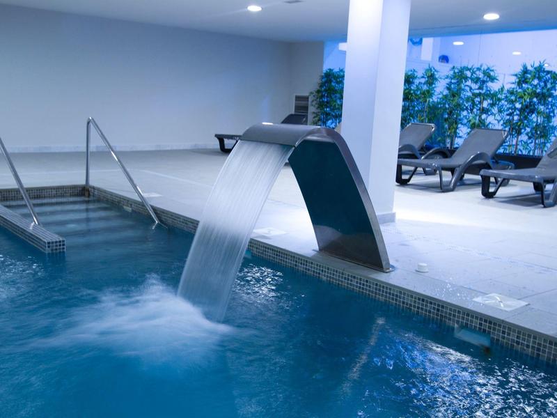 Piscina interna moderna con cascata e lettini in un'area benessere luminosa.