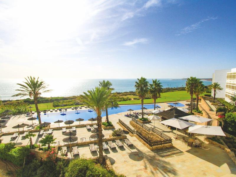 Piscina dell'hotel con lettini, palme e vista mare sotto un cielo limpido.