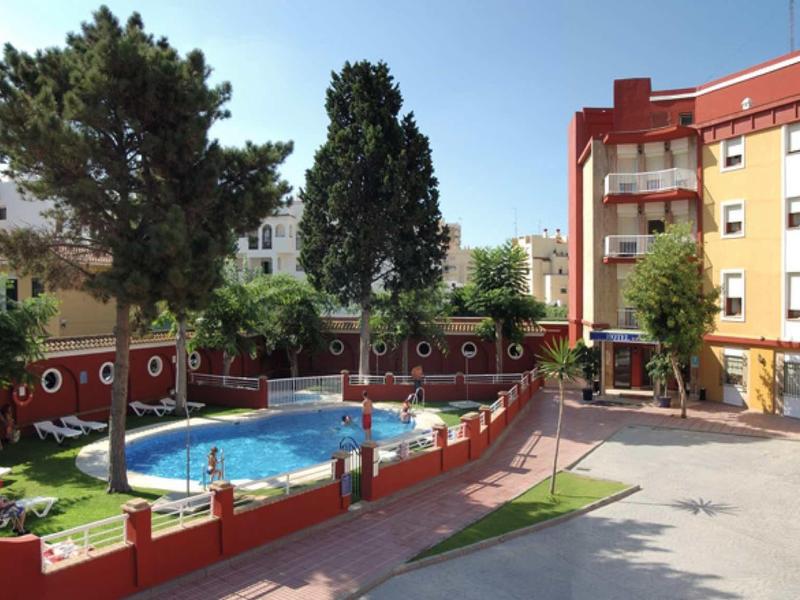 Hôtel avec piscine extérieure, entourée de clôtures rouges et de bâtiments sous un ciel bleu.
