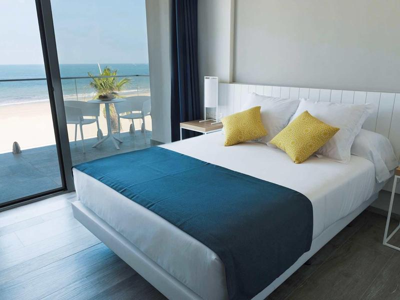 Chambre d'hôtel moderne avec vue sur la mer, grand lit, accents bleus et jaunes, et balcon.