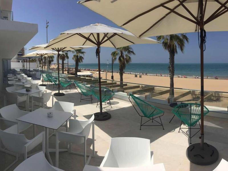 Vue d'une terrasse d'hôtel avec tables, parasols, piscine et palmiers au bord de la plage.