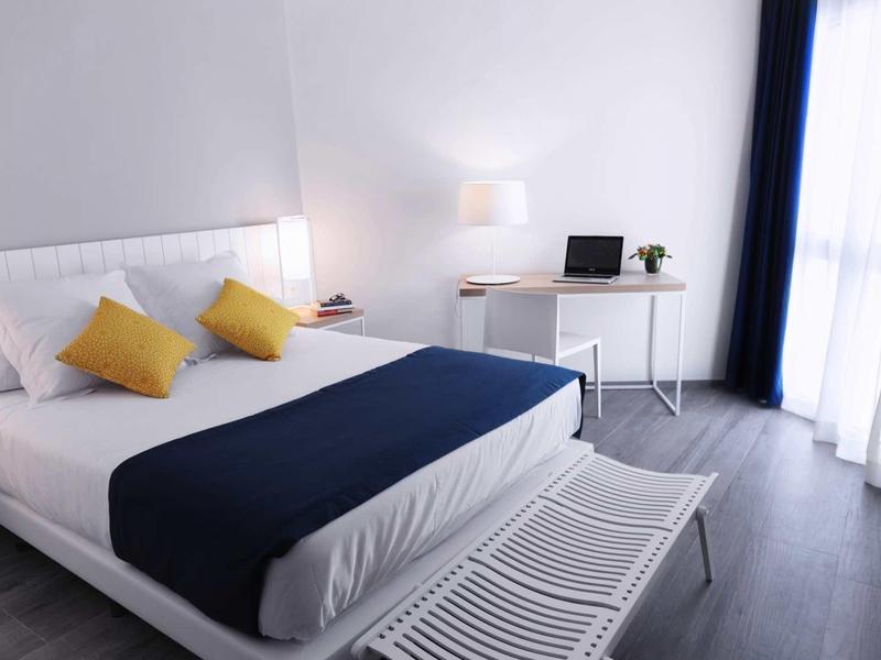 Chambre d'hôtel moderne avec lit double, murs blancs et grande fenêtre avec rideaux légers.