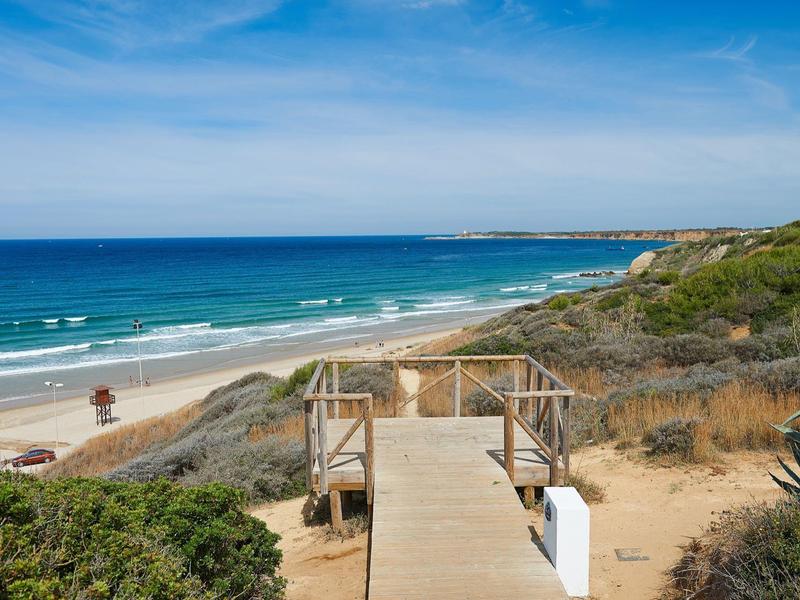 Wooden walkway leads to a long sandy beach with blue sea under clear sky.