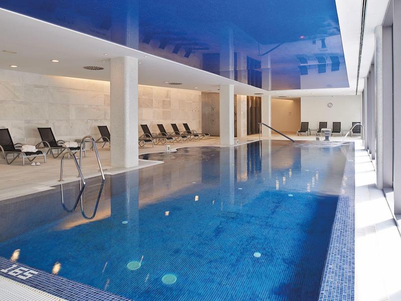 Modern indoor pool with blue ceiling and lounge chairs along the side.