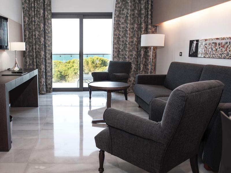 Modern hotel room with gray armchairs, sofa, and sea view through large windows.