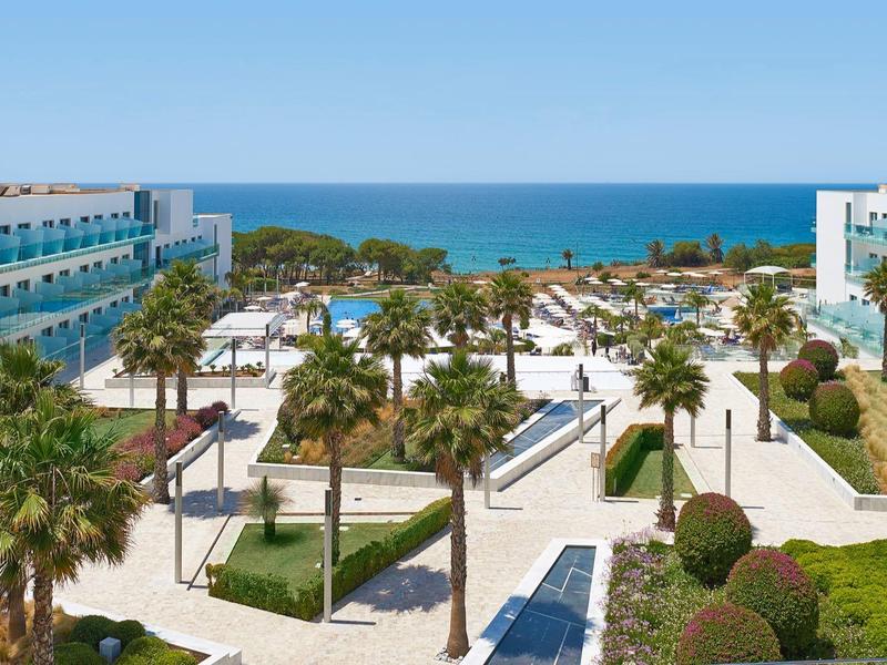 View of hotel complex with garden, palm-lined paths, and sea in the background on a sunny day