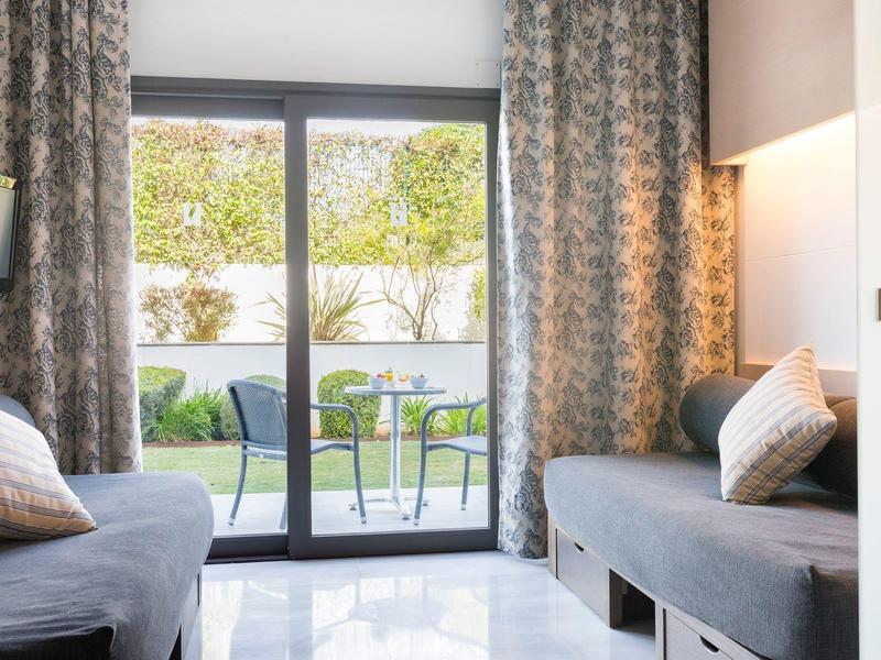 Bright hotel room with two sofas, cushions, and access to a terrace with garden furniture.