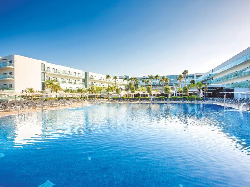 Large hotel pool with sun loungers and modern buildings under clear sky