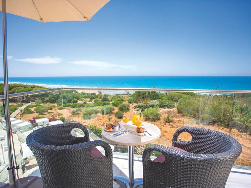 Two wicker chairs with a small round table on a balcony overlooking the sea with a sunshade.