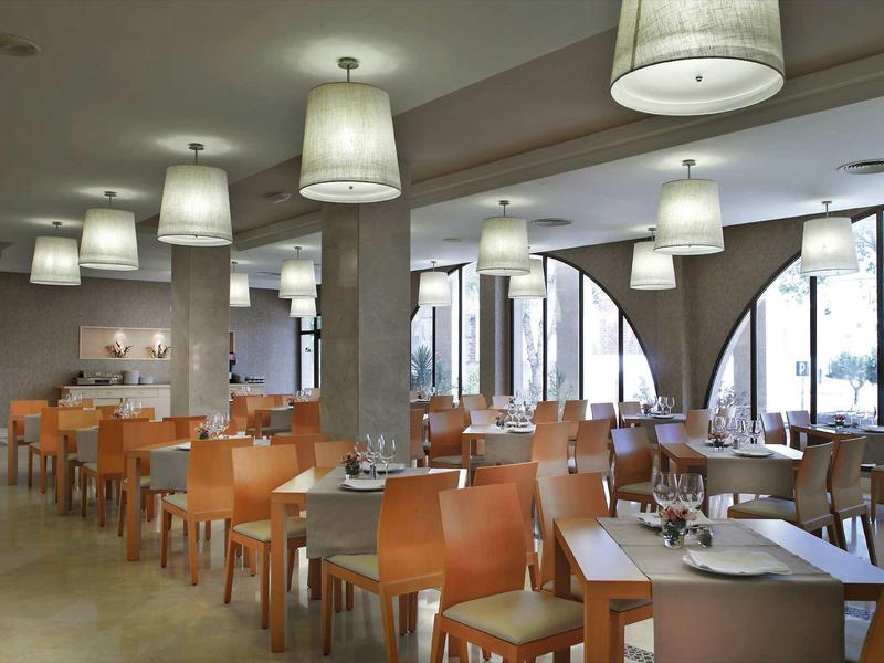 Moderner Speiseraum mit orangenen Stühlen, weißen Tischen, großen Lampen und bodentiefen Fenstern.