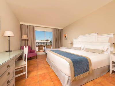 Lichte hotelkamer met groot bed, bureau, fauteuils en balkon met zeezicht.