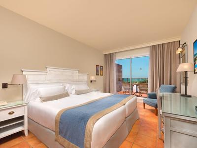 Lichte hotelkamer met tweepersoonsbed, balkon en zeezicht bij daglicht.