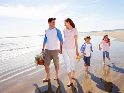 Familie met twee kinderen en picknickmand die blootsvoets langs het strand loopt.
