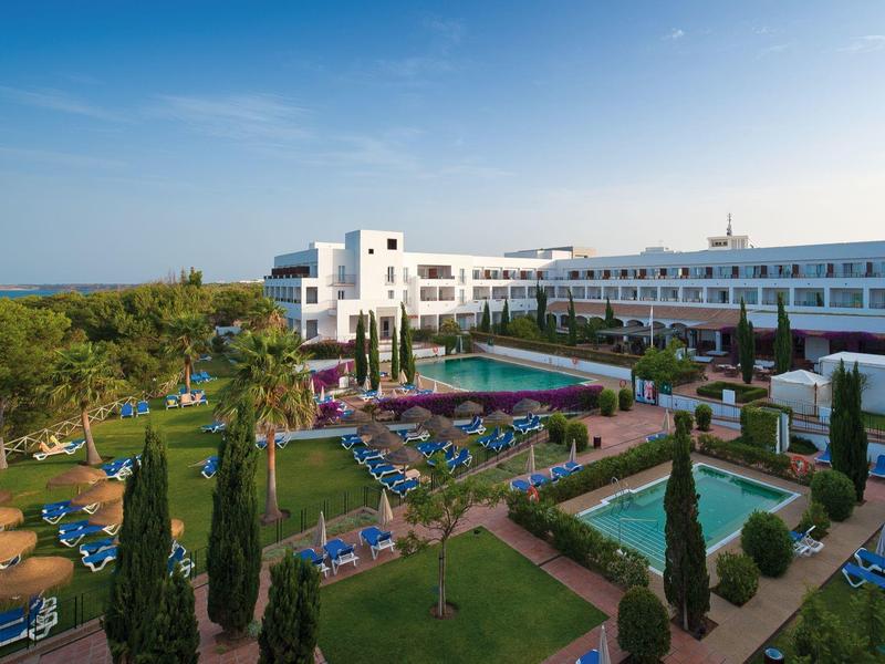 Groot hotel met zwembad, ligstoelen en verzorgde tuin onder een blauwe lucht.