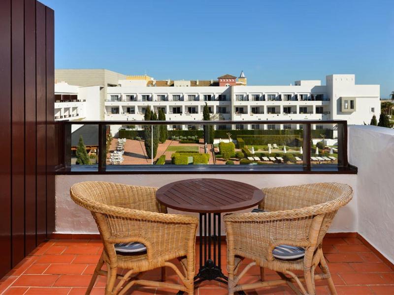 Balkon met tafel en twee rieten stoelen met uitzicht op moderne appartementen bij een blauwe lucht.