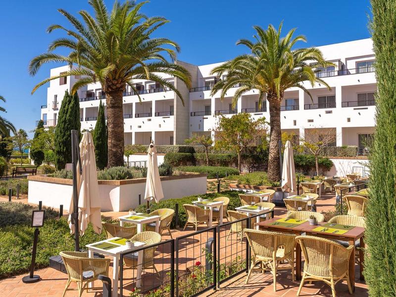 Buitengebied van een hotel met tafels, stoelen, parasols en palmbomen onder een heldere lucht.