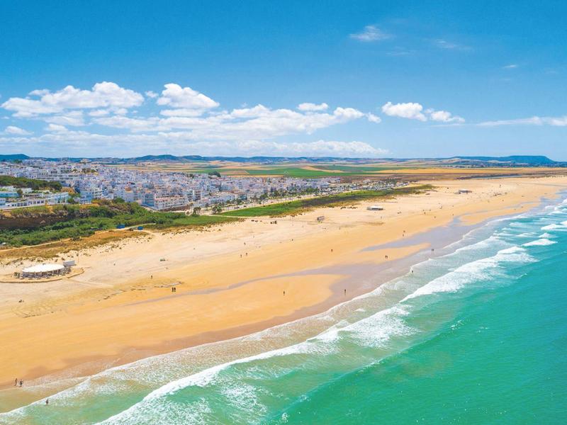 Uitgestrekt zandstrand met turquoise zee en stad op de achtergrond onder blauwe hemel.