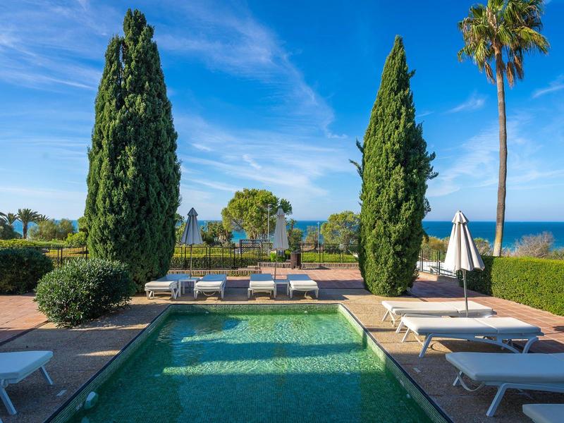 Uitzicht op een zwembad met ligstoelen, bomen en zee op de achtergrond bij heldere lucht.