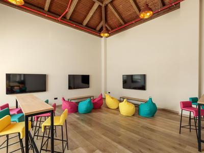 Nowoczesny hotelowy salon z drewnianą podłogą, telewizorami na ścianie i kolorowymi workami sako oraz krzesłami.