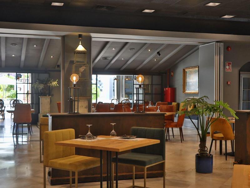 Gezellige hotel lounge met houten tafels, mosterdgele stoelen, planten en zachte verlichting.