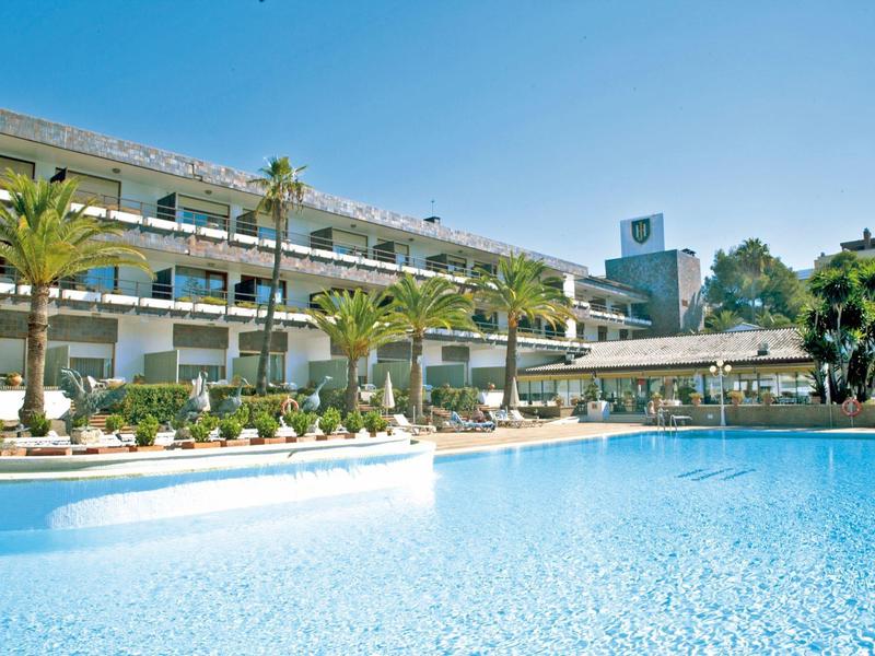 Hotel met buitenzwembad, omgeven door palmbomen onder een heldere blauwe lucht.