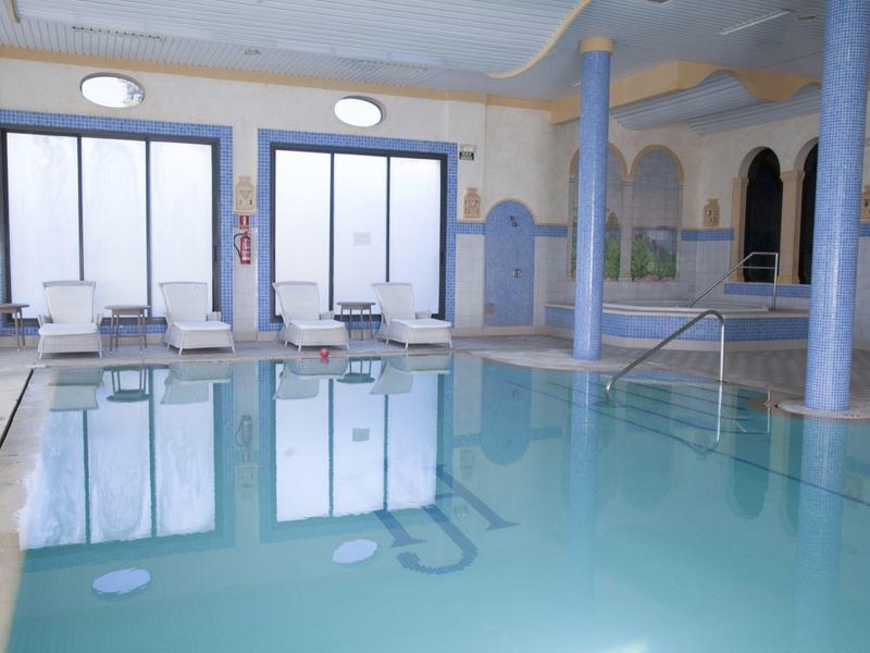 Binnenzwembad met blauwe zuilen, ligstoelen en grote ramen die licht binnenlaten.
