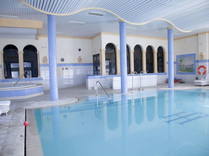 Overdekt hotelzwembad met zuilen, ligstoelen en betegelde muren onder golvend plafond.