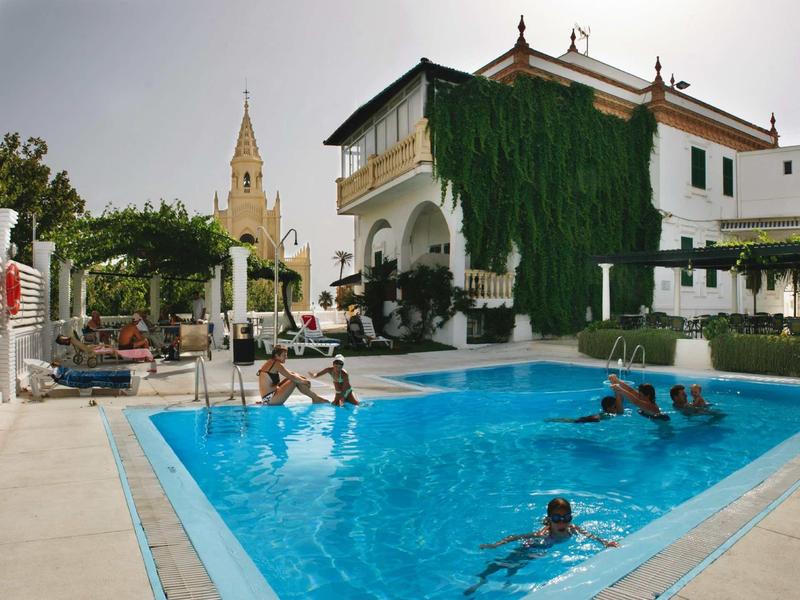 Piscina davanti a un hotel bianco con piante rampicanti e campanile sullo sfondo