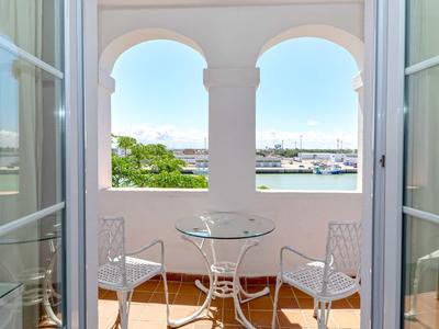 Balcony with white chairs and table overlooking a marina through arched windows.