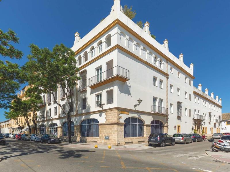 White multi-story building with balconies, located at a sunny street corner with parked cars.