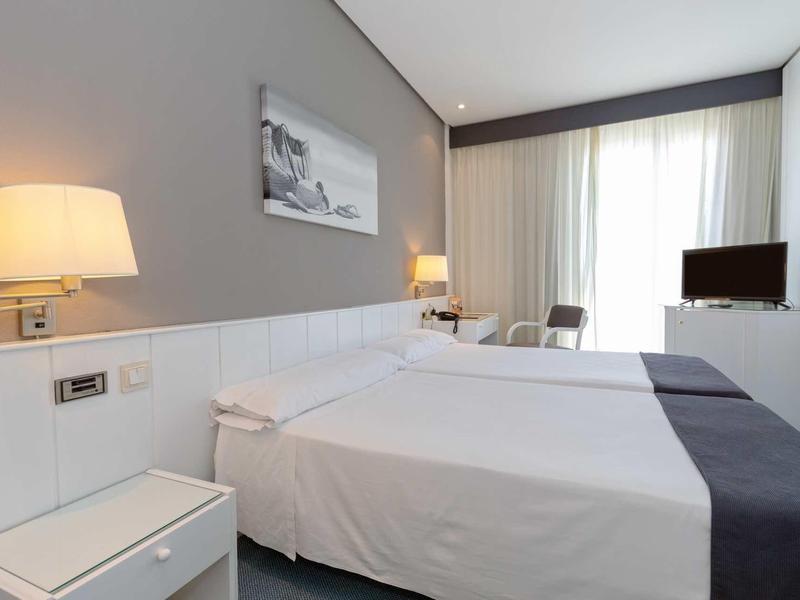 Bright hotel room with two single beds, white linens, bedside lamps, and a TV by the window.
