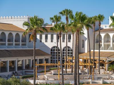 Exteriér bílého hotelu s palmami v popředí za slunečného počasí.