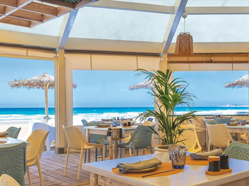 Příjemná restaurace u pláže s výhledem na moře a dřevěnou terasou pod skleněnou střechou.