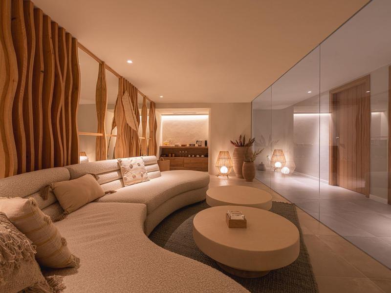 Moderní lounge s zakřivenou béžovou pohovkou, kulatými stoly, teplým osvětlením a vzorovanou dřevěnou stěnou.