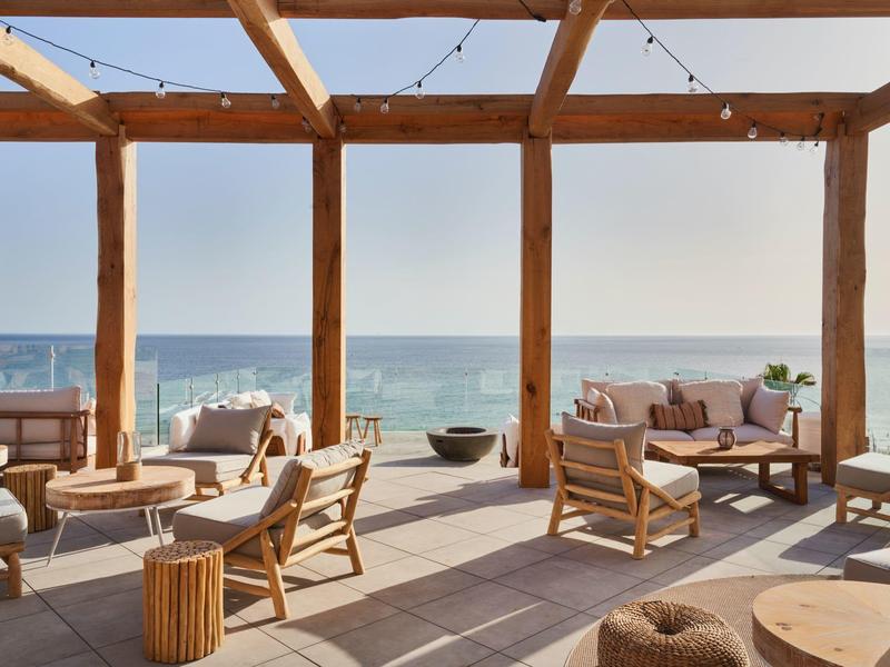Moderní venkovní posezení s pohodlným nábytkem a výhledem na moře pod dřevěnou pergolou.