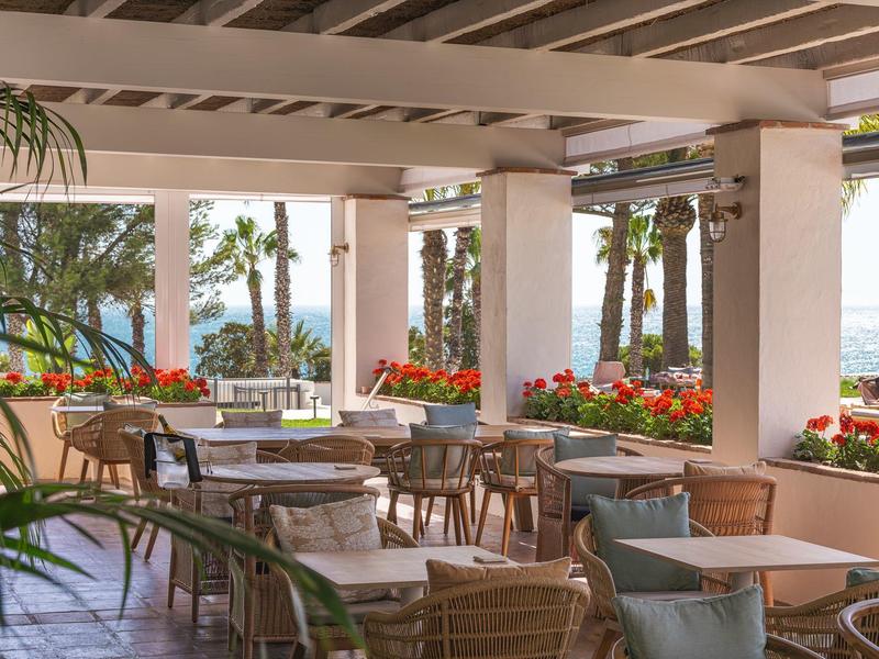 Venkovní hotelová terasa s dřevěným nábytkem, rostlinami a výhledem na moře.