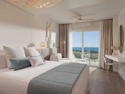 Hell eingerichtetes Hotelzimmer mit großem Bett und Meerblick durch bodentiefe Fenster.