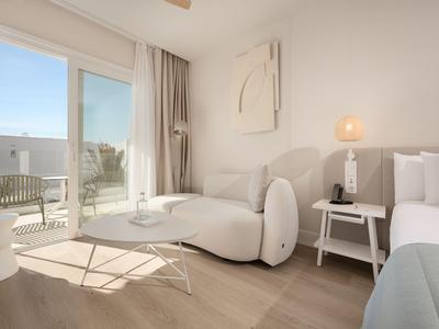 Helles Hotelzimmer mit modernem weißen Sofa, Tisch und Zugang zu Balkon bei Tageslicht.